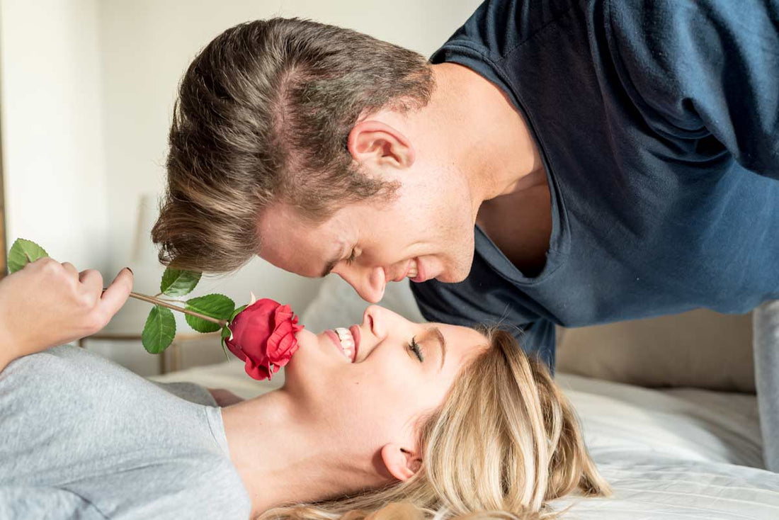 Romantic couple sharing a playful moment with a red rose while smiling on the bed — intimate and flirty relationship scene.