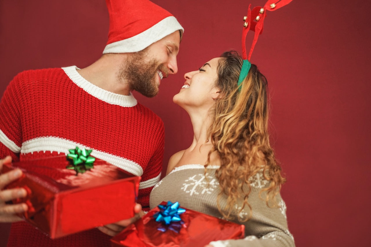 A smiling couple exchanging wrapped Christmas gifts in festive sweaters and Santa hats — perfect image for holiday gift ideas, Christmas sales, and seasonal product collections.